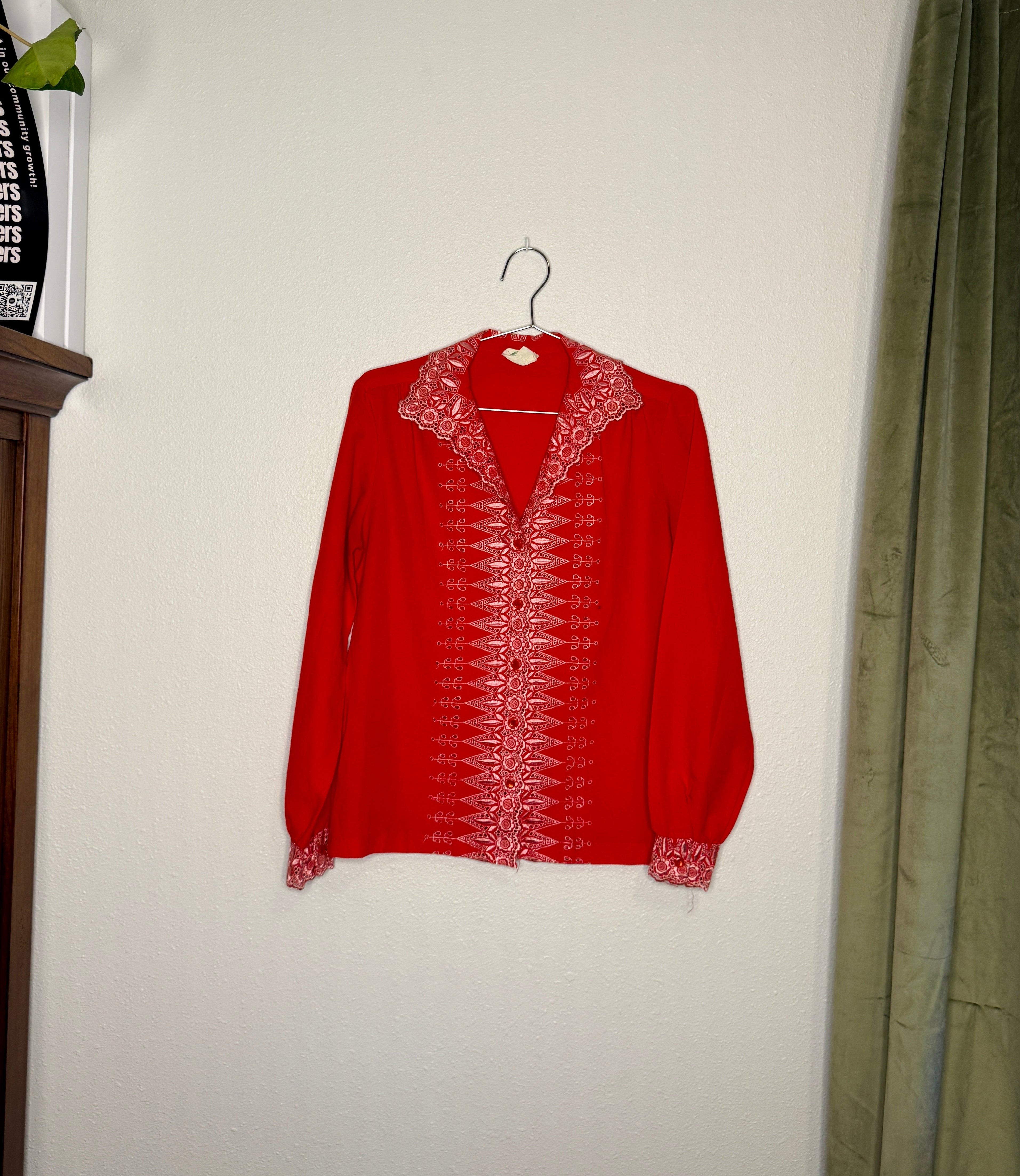 Red embroidered blouse on a hanger against a white wall with a curtain to the right.